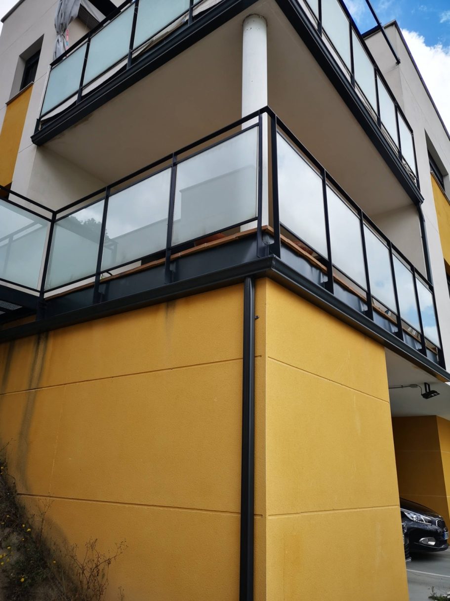 Balcón con barandilla de vidrio en un edificio de color amarillo y cielo nublado.