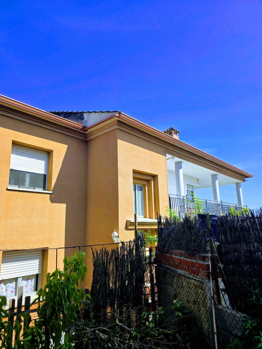 Casa de dos plantas con paredes amarillas y jardín, bajo un cielo azul claro.