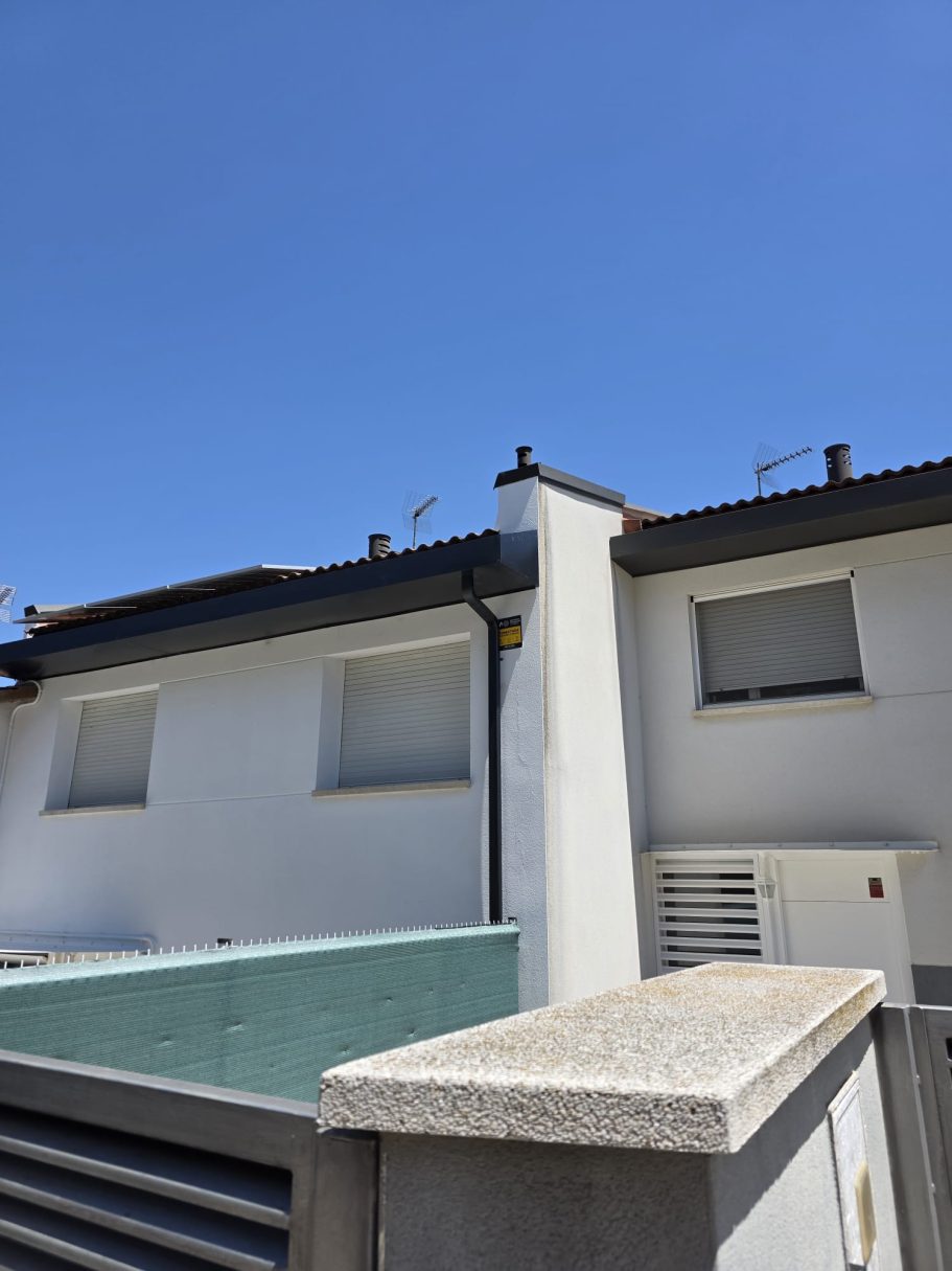 Edificio de dos plantas con tejado negro, cielo claro y un muro de piedra en primer plano.