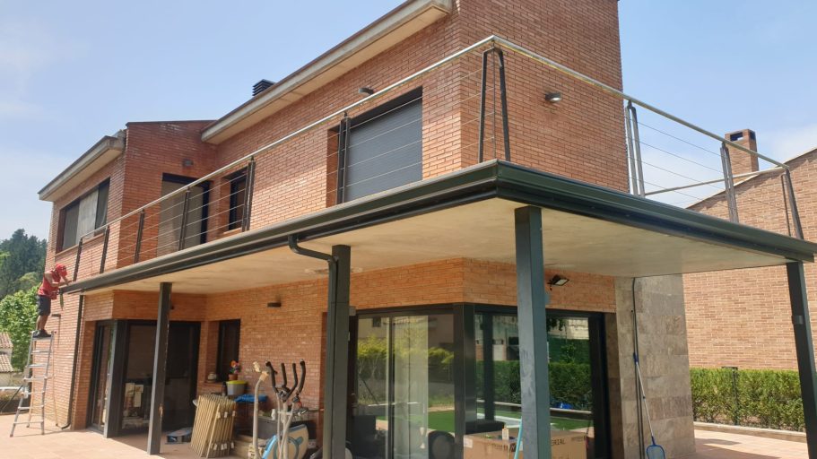 Casa de dos plantas de ladrillo, con balcón y ventanas amplias, rodeada de jardín.