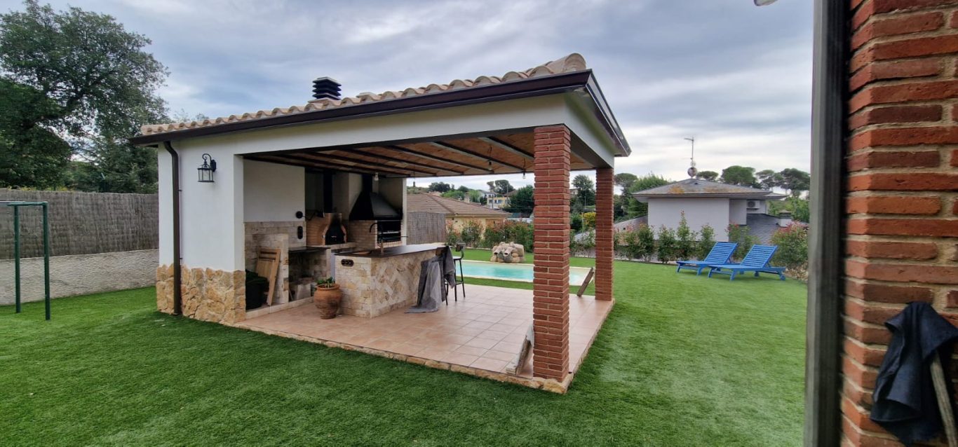 Casa de campo con porche, cocina exterior y área de descanso junto a la piscina.