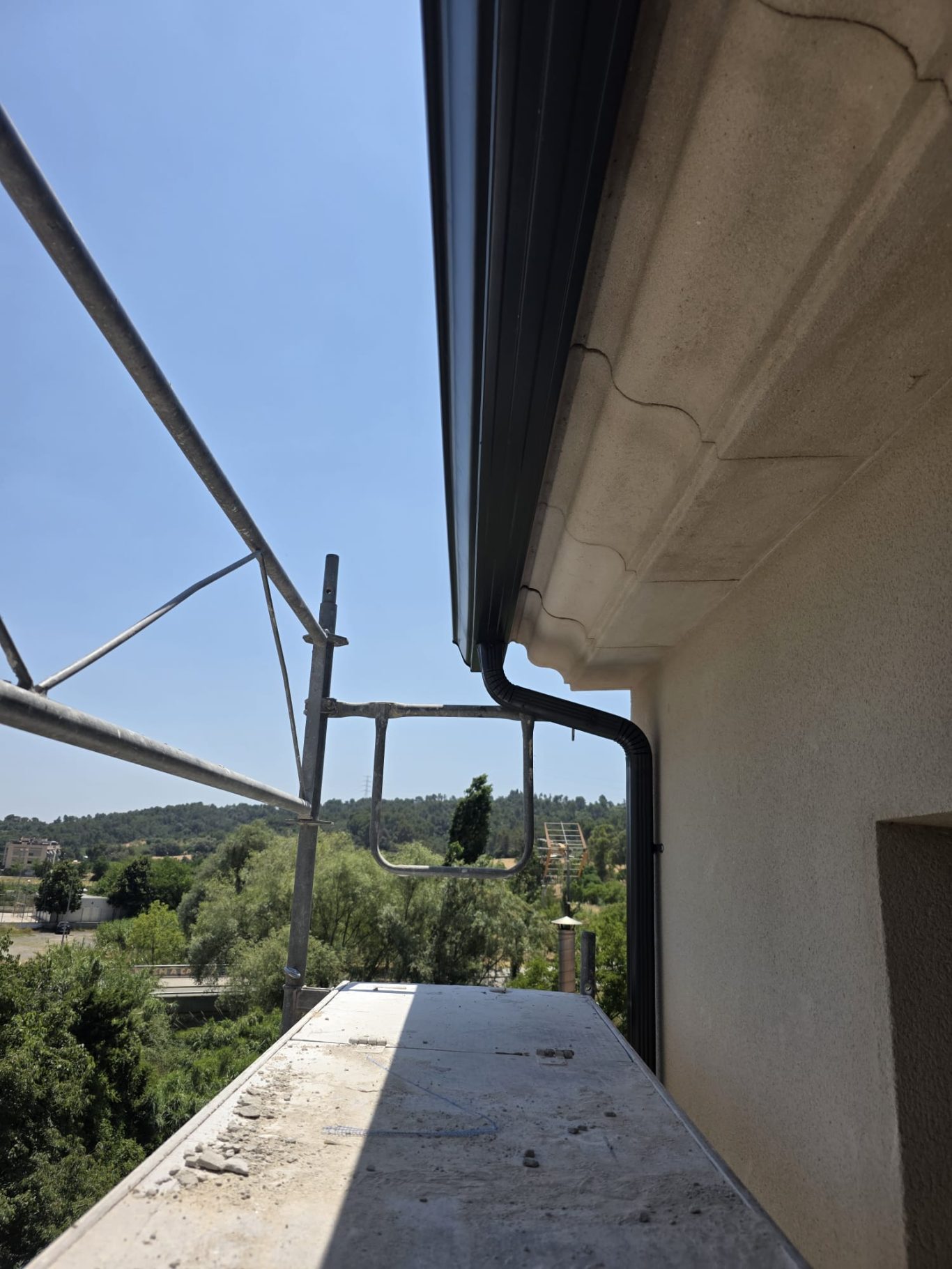 Vista de un balcón con andamios y paisaje arbolado al fondo bajo un cielo despejado.