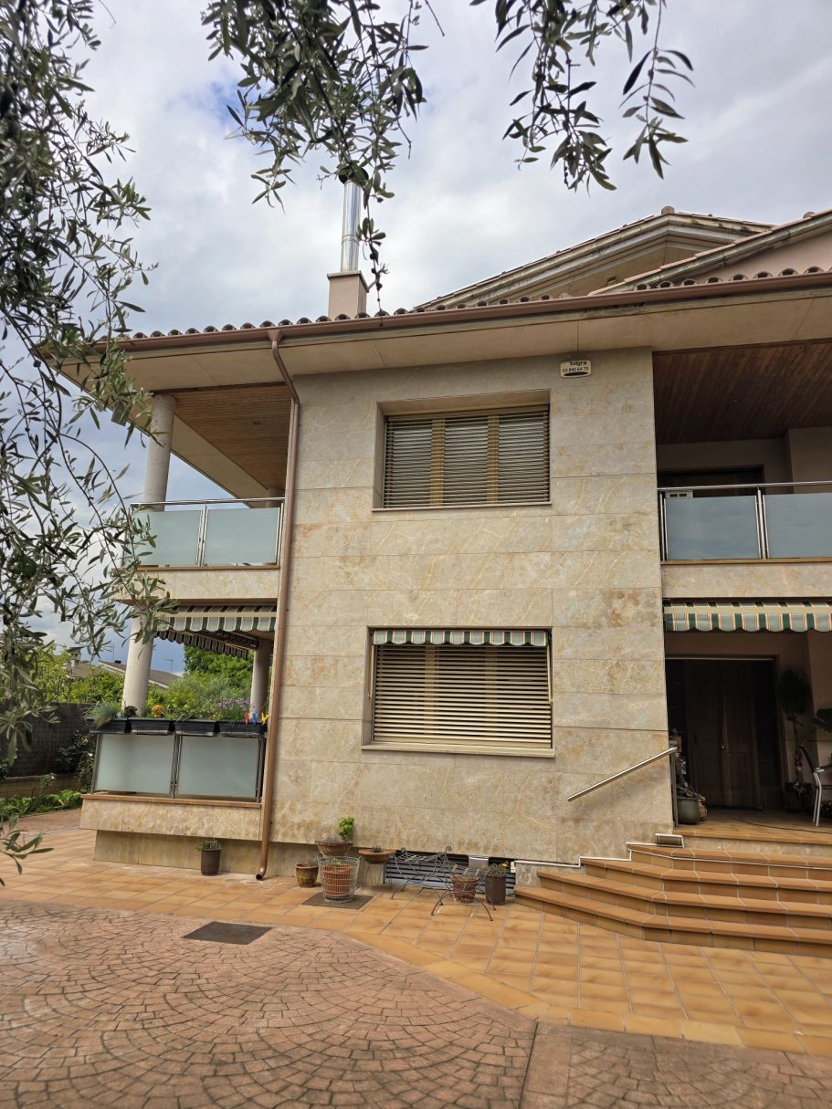 Casa de dos plantas con balcón, acceso a escaleras y vegetación cercana.